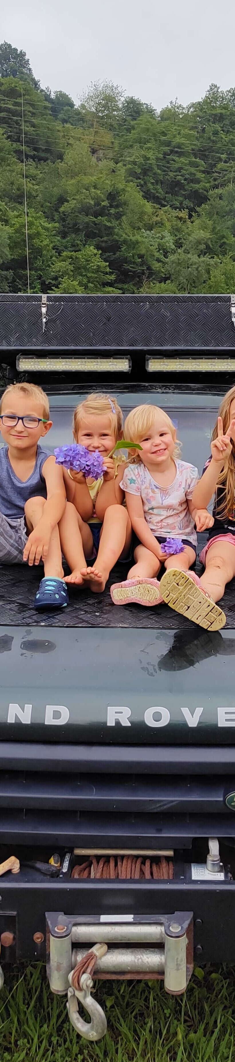 Czworo dzieci siedzących na masce samochodu terenowego zaparkowanego na trawie