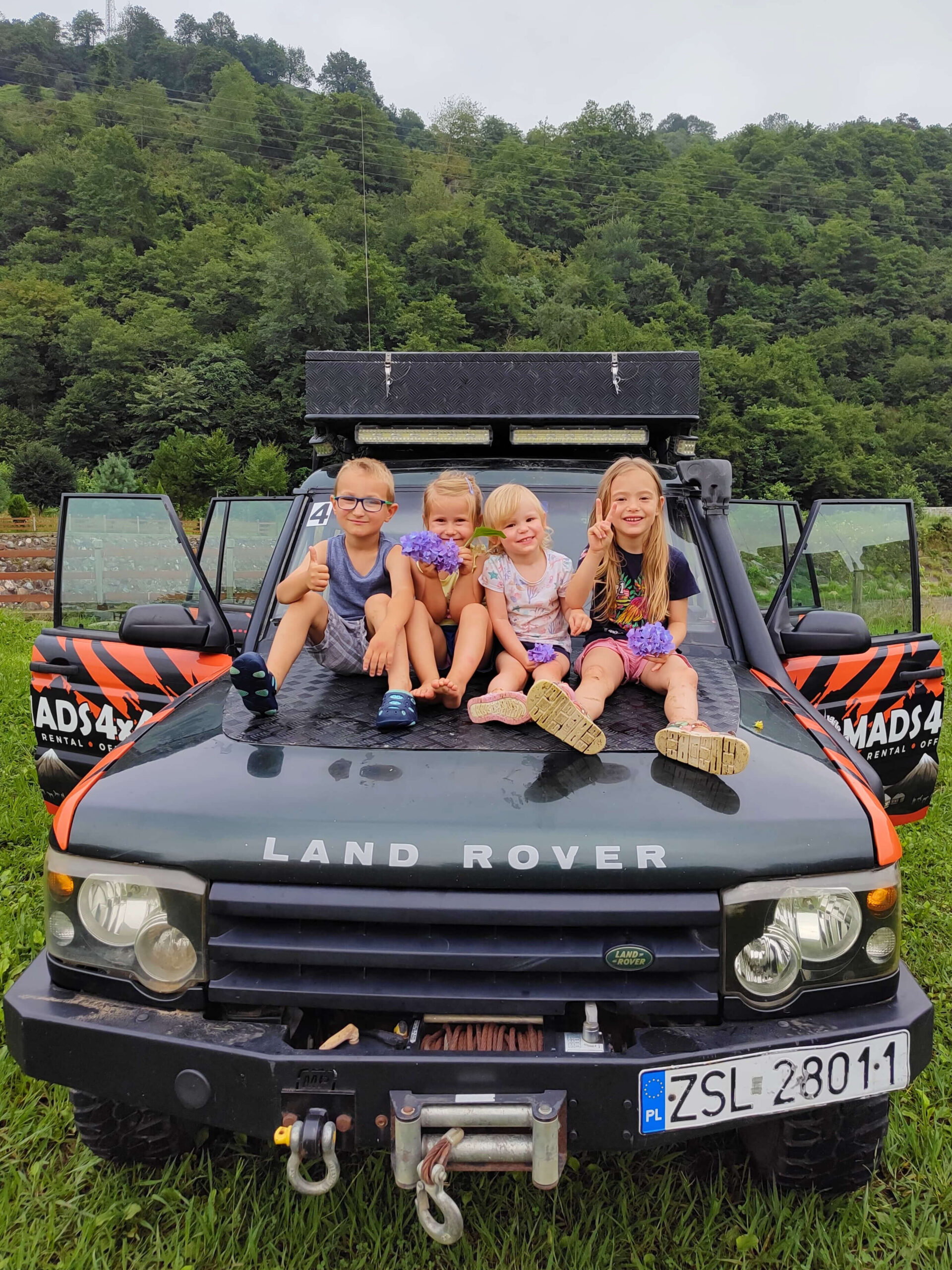 Czworo dzieci siedzących na masce samochodu terenowego zaparkowanego na trawie